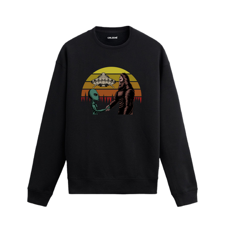 Uzay temalı siyah sweatshirt üzerinde uzaylı ve kocaayak figürlerinin el sıkıştığı, arkada UFO ve retro güneş tasarımı bulunan yaratıcı illüstrasyon.